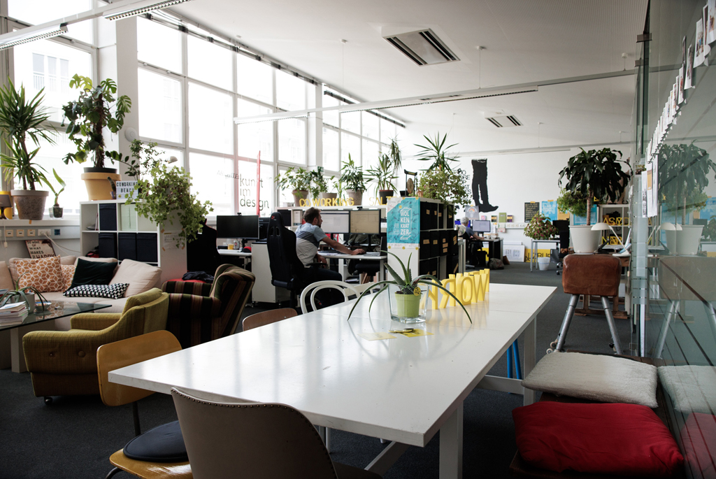 Hohes helles Großraumbüro mit großem Tisch im Vordergrund und mehren Coworking-Arbeitsplätzen im Hintergrund vor einer großen hellen Fensterfläche und Grünpflanzen.